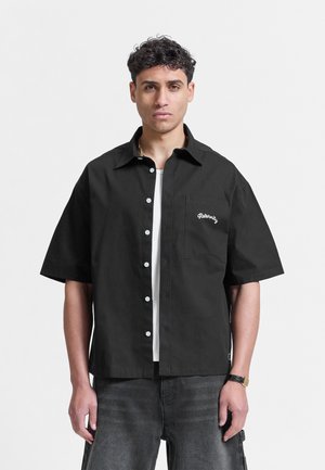 Man met kort krullend haar, gekleed in een zwart kort mouw knoopoverhemd over een wit T-shirt, staand tegen een effen lichte achtergrond.