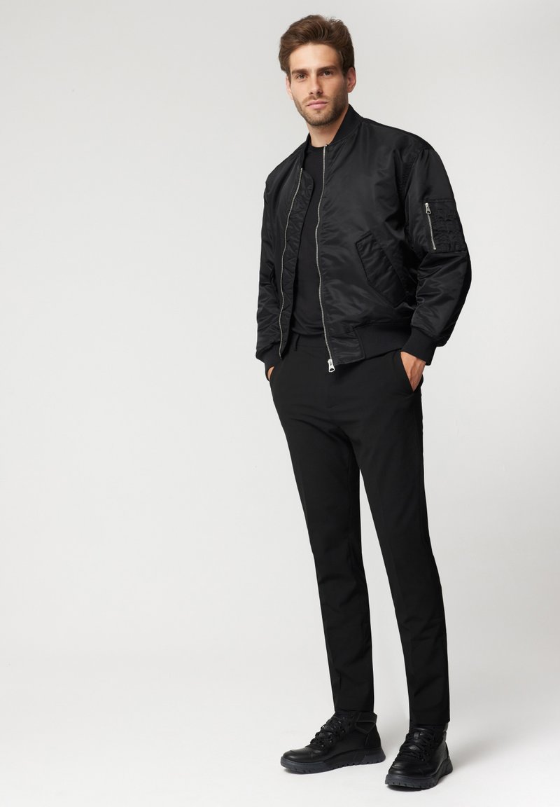 Czarna kurtka bomberka o gładkiej, błyszczącej fakturze, z zapięciem na zamek, kieszeniami po bokach i ściągaczami na mankietach, w połączeniu z czarnymi spodniami slim-fit i sneakersami.