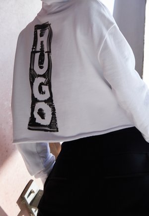 Persoon draagt een witte cropped hoodie met dikke zwarte verticale "VOGUE" tekst op de achterkant en een zwarte broek, staand binnen bij een raam.