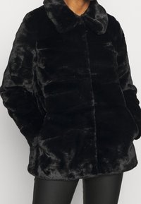 Manteau en fausse fourrure noire avec col, présentant une texture douce et pelucheuse, fermeture avant à boutons, et poches latérales. La coupe est ample et décontractée.