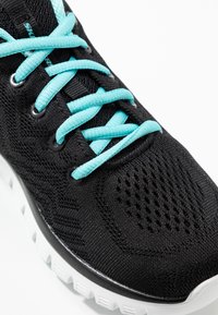 Zapatilla deportiva de malla negra con cordones azul claro y suela blanca, mostrando un primer plano de los detalles superiores y laterales.