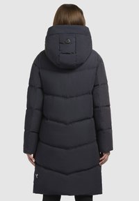 Dunkel pufferjakke med hætte, der har chevron-syninger, et rundt logo på ryggen og en mellemlang længde. Glat tekstur.