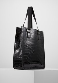 Sac fourre-tout noir en matériau brillant et texturé avec un motif peau de crocodile, doté de deux poignées solides et d'une forme rectangulaire.