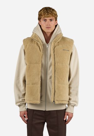 Beige Fleeceweste mit Reißverschluss, Seitentaschen und einer weichen, strukturierten Oberfläche, getragen über einem beigen Kapuzenpullover und braunen Hosen.