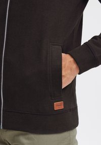 Svart zip-up hettegenser med ribbestrikkede mansjetter og liv, med en frontlomme og et teksturert lærlogo-patch. Stoffet ser mykt ut.