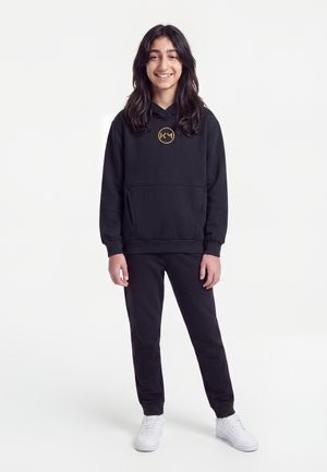 Une jeune personne souriante porte un sweat à capuche noir avec un logo doré, un pantalon noir et des baskets blanches sur un fond blanc uni.