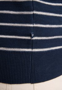 Pulover alb și albastru cu dungi, cu tiv din tricot și fante laterale, având un mic etichetă cu logo aproape de cusătură. Material moale din tricot.