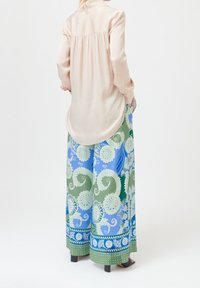 Vrouw in een lichtroze blouse met lange mouwen en een gathered achterkant, gecombineerd met wijde broek in blauwe en groene ingewikkelde bloemenpatronen.