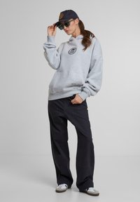 Hanorac gri cu glugă și logo, asociat cu pantaloni largi negri. Accesoriile includ o șapcă închisă și ochelari de soare. Încălțăminte lejeră este prezentă.