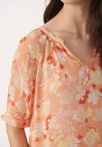 Blouse florale pêche avec un col en V, des manches courtes retroussées et un tissu léger, légèrement transparent. Présente un motif floral en blanc et rouge.