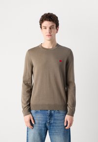 Langermet genser i olivengrønn strikk, med rund halslinning og en liten rød logo-patch på brystet. Kombinert med blå jeans.
