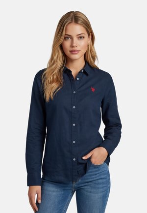 Junge Frau trägt ein marineblaues Hemd mit rotem besticktem Logo und blaue Jeans, steht mit einer Hand in der Hosentasche.