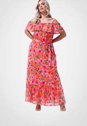 Frau trägt ein langes, schulterfreies oranges Kleid mit pinkem Blumenmuster und einem Bindegürtel, steht mit Händen in die Hüften gestützt vor weißem Hintergrund.