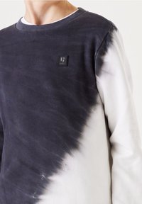 Grauer und weißer Batik-Sweatshirt mit Rundhalsausschnitt, weichem Material und einem quadratischen Logopatch auf der Brust. Verschwommener Farbverlauf.