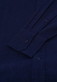 Tmavo modrá košela s dlhým rukávom a zapínaním na gombíky na manžetách a prednej strane, poskladaná a položená na tmavom pozadí.