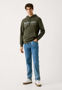 Sudadera verde con el logo de Red Bull Racing, bolsillo frontal y capucha con cordón. Combinada con vaqueros azules y zapatillas blancas con suela de goma.