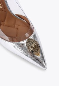 Pompe argentée à talons aiguille avec des côtés transparents et un accent en plumes dorées à l'avant, ornée de pierres noires sur une surface texturée.