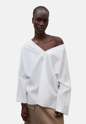 Mørkhudet kvinde med fletninger, iført en overdimensioneret hvid skulderfri skjorte og beige nederdel mod en ensfarvet baggrund.