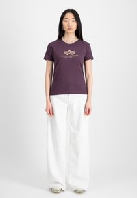 T-shirt violet à manches courtes avec un imprimé logo doré, accompagné d'un large pantalon blanc. Texture en coton lisse avec une coupe ajustée.