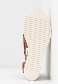 Chaussure en cuir marron avec un design perforé, affichant une semelle en caoutchouc rayée blanche. L'arrière est courbé, avec une surface texturée.