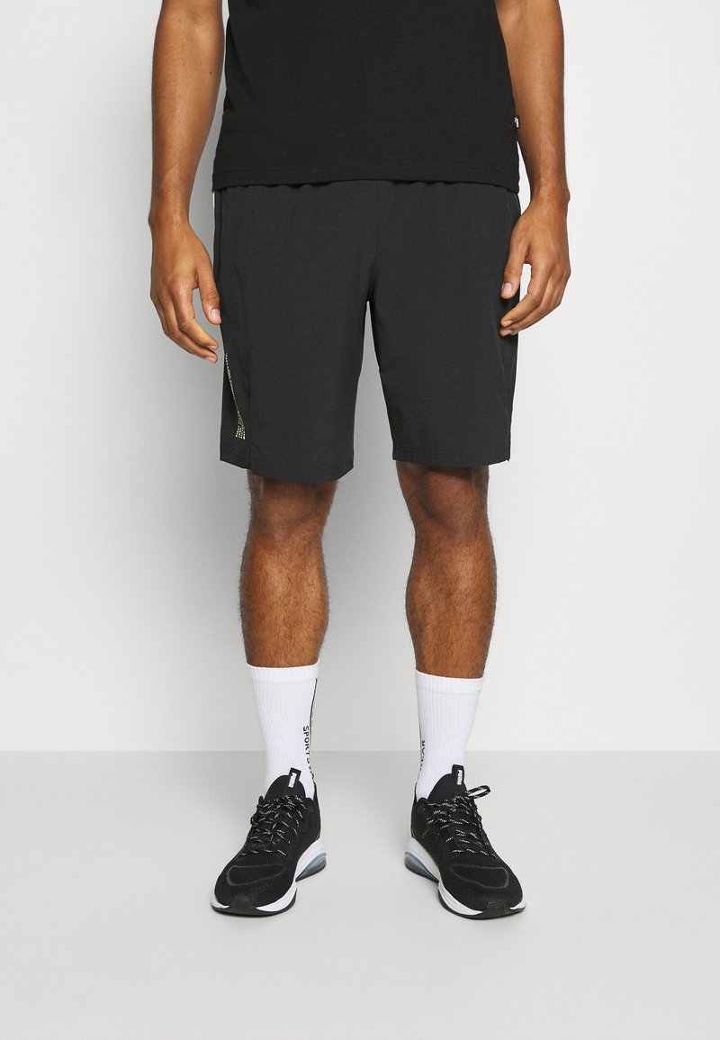 Homme debout portant un short de sport noir, des baskets noires avec des semelles blanches, et des chaussettes blanches à mi-mollet avec du texte noir sur un fond uni.