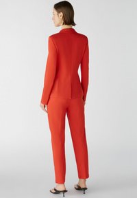 Roter, taillierter Anzug mit einem maßgeschneiderten Blazer und schmal zulaufenden Hosen. Glatte Textur und strukturiertes Design. Tragen mit schwarzen High Heels.