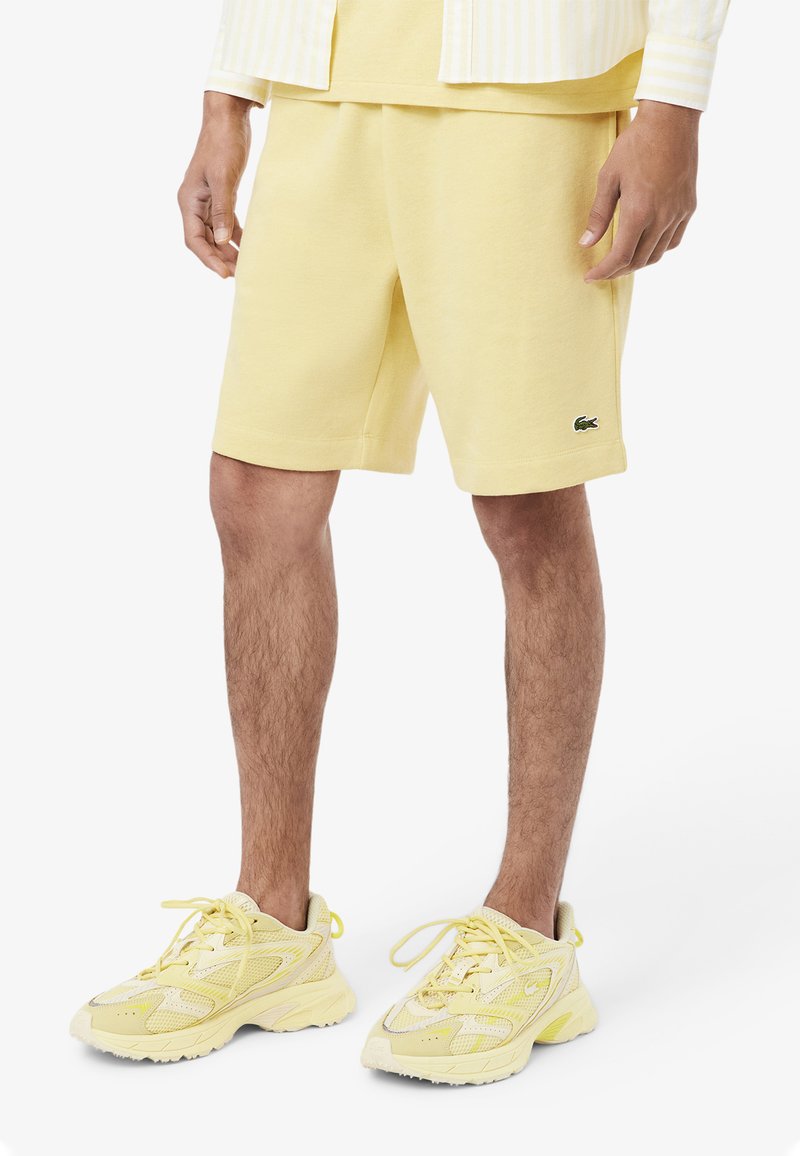 Shorts di cotone gialli con tasche e logo ricamato; abbinati a sneakers gialle con dettagli in rete e suole in gomma.