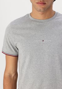 Harmaa puuvillainen t-paita, jossa on pyöreä pääntie, lyhyet hihat punaisin ja sinisin raidoin, sekä keskellä oleva logo.