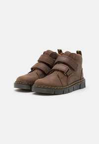 Botas de cuero marrón con dos correas de Velcro ajustables, punta redonda, suela de goma y costura amarilla a lo largo del borde. Parte superior texturizada, altura hasta la mitad de la pantorrilla.