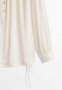 Blouse crème à manches longues avec poignets boutonnés, deux boutons près de l'encolure et cordon ajustable à l'ourlet.