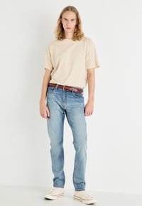 Beige Baumwoll-T-Shirt, hellblaue Denim-Jeans, geflochtener brauner Ledergürtel, weiße Sneakers mit roten Akzenten, vor einem einfarbigen Hintergrund stehend.
