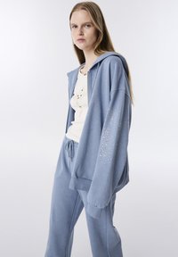 Felpa azzurra chiara con strass decorativi, abbinata a pantaloni della tuta coordinati. Il tessuto sembra morbido con una vestibilità comoda e vita regolabile con cordoncino.
