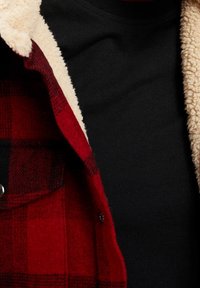 Rood en zwart geruite jas met beige fleece voering, gedragen over een zwarte top, waarbij de details van de bovenste borst en kraag zichtbaar zijn.