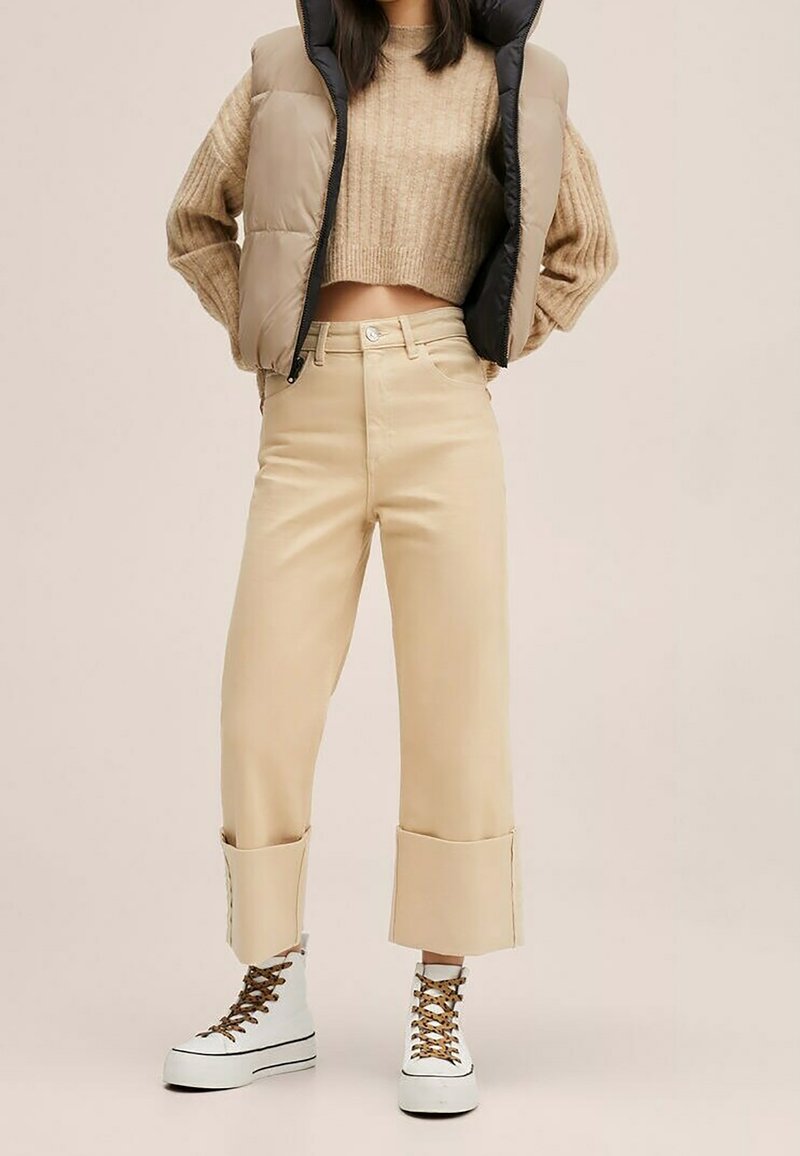 La femme porte un pull court beige, un pantalon large beige taille haute avec de grands revers, une veste matelassée, et des bottines blanches à plateforme à lacets.