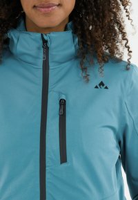 Tiefblaue Jacke mit hohem Kragen, durchgehendem Reißverschluss, schwarzen Reißverschlussakzenten und einem kleinen schwarzen Logo auf der oberen linken Seite. Glatte Textur.