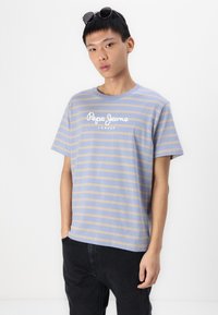 T-shirt en coton rayé bleu et beige avec col rond, arborant le logo "Pepe Jeans London". Assorti à un pantalon en denim noir et des lunettes de soleil.