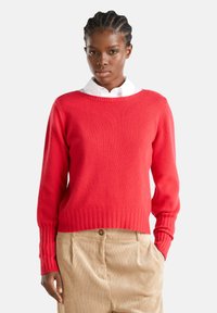Joven con el cabello trenzado, vistiendo un suéter rojo sobre una camisa blanca con cuello y pantalones de pana beige, con una mano en el bolsillo.