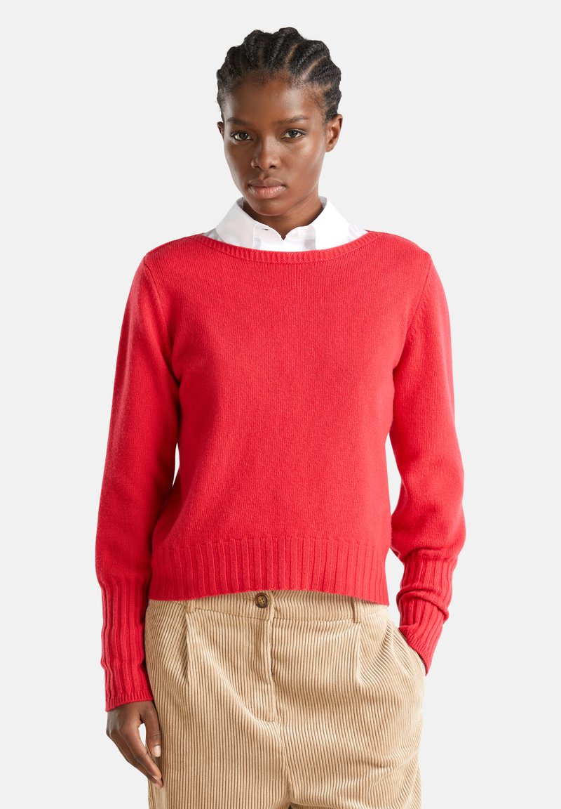 Joven con el cabello trenzado, vistiendo un suéter rojo sobre una camisa blanca con cuello y pantalones de pana beige, con una mano en el bolsillo.