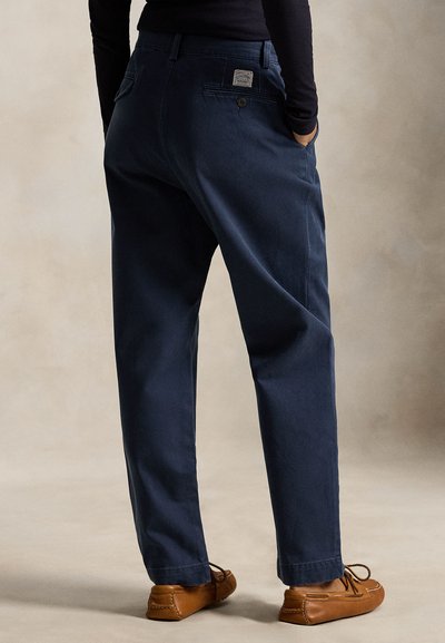 Persoon die donkerblauwe, ruimvallende broek met achterzakken en bruine leren loafers draagt, staand met handen in de zakken.
