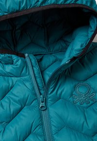 Tyrkysová prošívaná puffer bunda s kapucí, vybavená předním zipem a vyšitým logem na levé straně. Hladká textura, diamantový vzor.