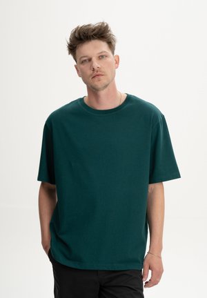 Jonge man met kort, warrig haar, gekleed in een loszittend donker groen T-shirt en zwarte broek, tegen een effen lichte achtergrond.