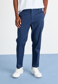 Mørkeblå bukser med lige snit, lavet af et glat stof. Har standardlommer og knaplukning i taljen. Parret med hvide sneakers.