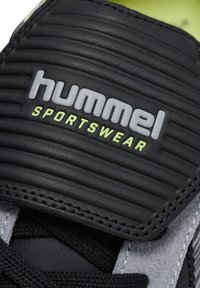 Close-up of black sports shoe tongue with textured surface, showing "hummel SPORTSWEAR" logo in gray and neon yellow letters.