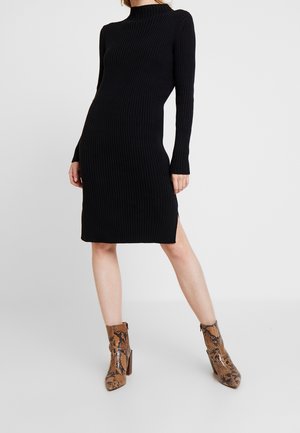 Frau trägt ein schwarzes, geripptes Strick-Midikleid mit langen Ärmeln, kombiniert mit braunen, hochhackigen Stiefeletten aus Schlangenleder, steht vor einem weißen Hintergrund.