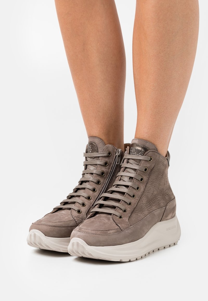 Wysokie skórzane trampki w kolorze taupe z bocznymi suwakami i grubymi białymi podeszwami, noszone na gołych nogach na białym tle.