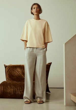 Pastelovo žltá nadrozmerná mikina, svetlošedé široké nohavice a béžové sandále na nízkom podpätku na osobe, na neutrálnom pozadí.