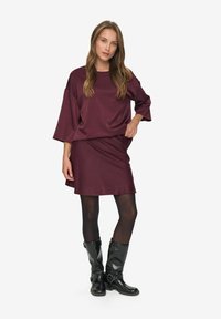 Femme portant un haut ample maroon et une jupe assortie, des collants noirs et des bottes noires mi-mollet, se tenant avec une main sur la hanche.