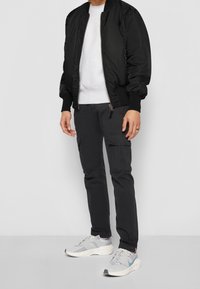 Personne portant une veste bomber noire, un sweat-shirt blanc, un pantalon cargo noir et des baskets gris clair avec des semelles mouchetées, debout devant un fond uni.