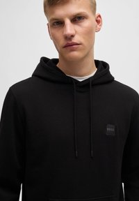 Svart hoodie i bomullsblandning med en känguruficka, dragsko på huvan och en liten fyrkantig logotyp i svart på bröstet.