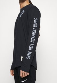 Czarna koszula z długim rękawem z logo na piersi oraz napisem "TEN SAM PIEKŁO INNE DIABŁY" na rękawie w pogrubionej czcionce. Materiał bawełniany.